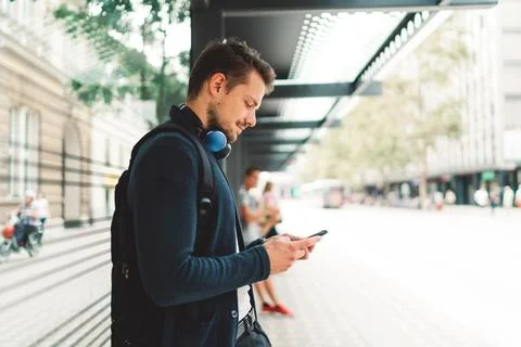 Side view of young man looking down at his phone while waiting for his bus to Stock Photos