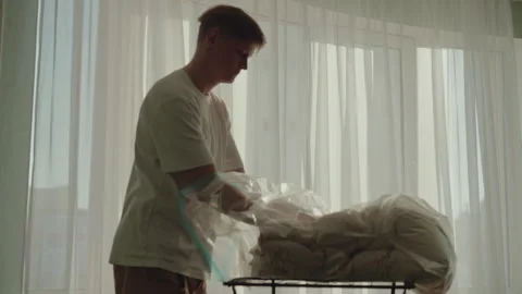 Side view of young man packing soft blanket for storage using vacuum cleaner and Stock Footage 305512537