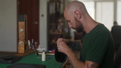 Side view of young professional leather craftsman sits at work table and makes Stock Footage 256296073
