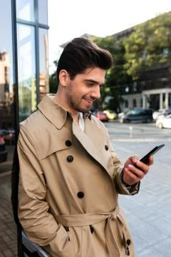 Side view of young smiling man in trench coat happily using cellphone on street Stock Photos