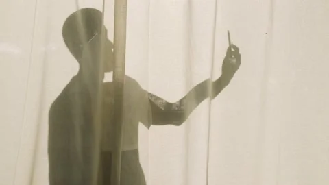 Silhouette of a bold man standing behind curtains with her smartphone Stock Footage 157284505