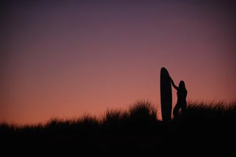 Silhouette Of Female Surfer Standing And Holding Surfboard Across Dunes Against Stock Photos