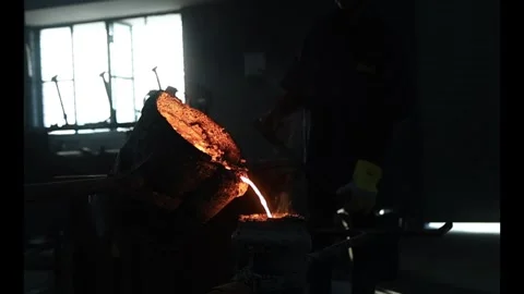 Silhouette of foundry worker pouring molten orange metal from a heavy crucible Stock Footage 337912234