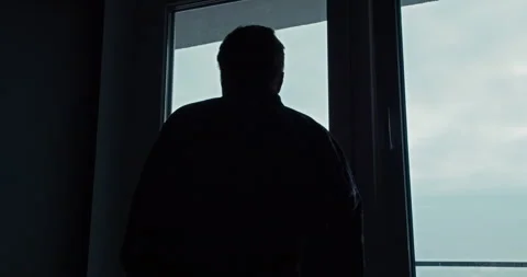 Silhouette of a guy looking out a blue window while standing in a dark gray Stock Footage 305790112