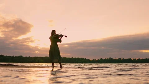 Silhouette of a lone violinist performing on a tranquil shoreline at sunset, Stock Footage 340212677