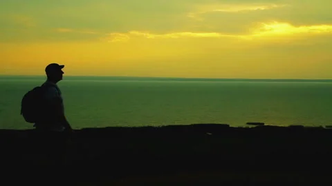 Silhouette of a man with a backpack behind his back on the edge of a hill at Stock Footage 95016034