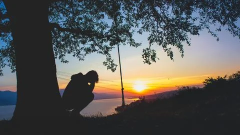 Silhouette of a man crouching under a tree during a dramatic sunset. Stock Photos
