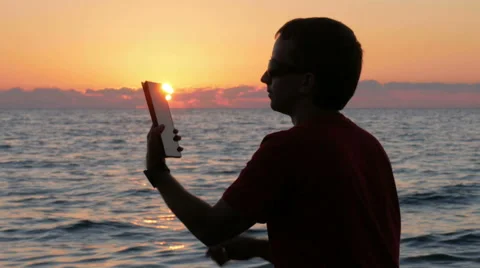 Silhouette man with digital tablet in hands at sunset beach. The sun is almost Stock Footage 67729113