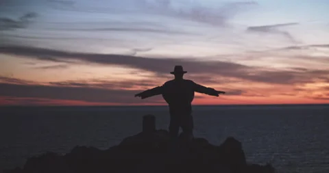 Silhouette of a man with a hat walking towards a cliff at dusk Stock Footage 203589756