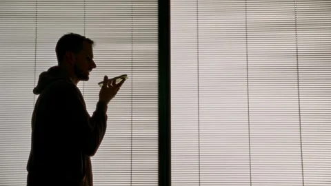 Silhouette of man recording voice message on smartphone standing next to window Stock Footage 311709953