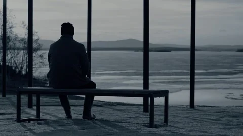 The silhouette of a man sitting alone on a bench by a lake with melting ice. Vídeo Stock 332498827