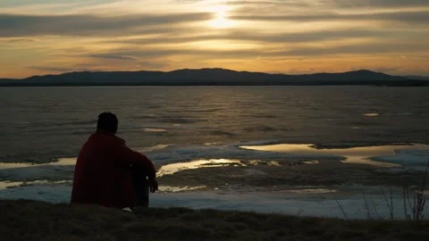 Silhouette of a man sitting alone on the shore of a lake at sunset. 스톡 동영상 332476492