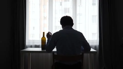Silhouette of a man sitting alone at a table, drinking alcohol from a bottle in Stock Footage 346250856