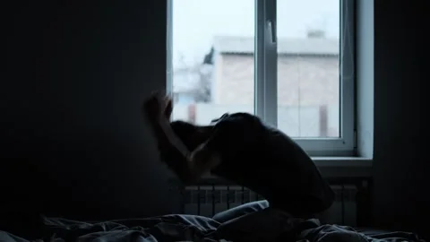 Silhouette of a man sitting on a bed by the window, emotionally hitting the bed  Video stock 296240427