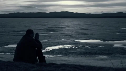 The silhouette of a sad man sitting alone on the shore of a lake. Video stock 332498822