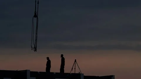 Silhouette two construction workers stand on rooftop of building with crane hook Stock Footage 323912333