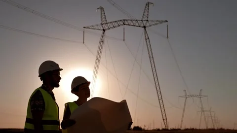 Silhouette of two electrical engineers in hard hats standing in field at sunset Stock Footage 201508225