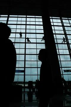 A silhouette of a worker cleaning windows hangs from a rope in the airport .. Stock Photos
