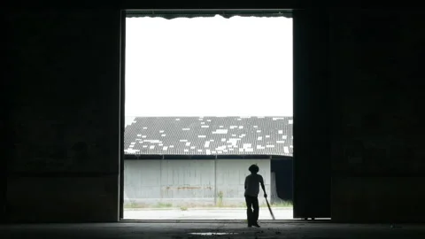Silhouette of a worker sweeping framed by a large industrial warehouse doorway Stock Footage 323067883