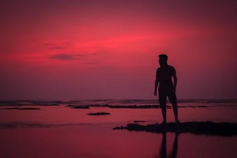 Silhouette of a young man standing in front of a colorful sunset at the beach Stock Photos