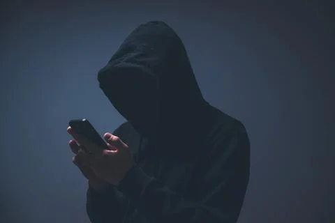 Silhouetted hacker in a dark hoodie using a smartphone, symbolizing hacking.. 库存照片