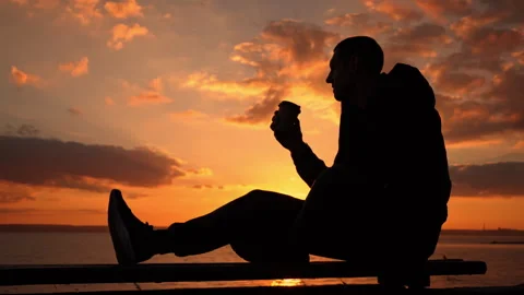 Silhouetted Man at Sunset Drinks Coffee on a Bench Stock Footage 256088103