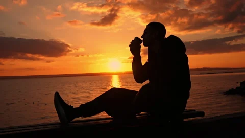 Silhouetted Man at Sunset Drinks Coffee on a Bench Stock Footage 256165426