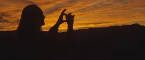 Silhouetted man watching sunset taking photos with mountains in background 库存影片 128499754
