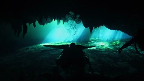 Silhouetted scuba divers exploring a sunlit underwater cave with stalactites and 스톡 동영상 331341365