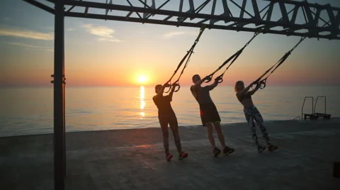 Silhouettes of sportive people training arms with trx at sunrise. Slow motion. Stock Footage