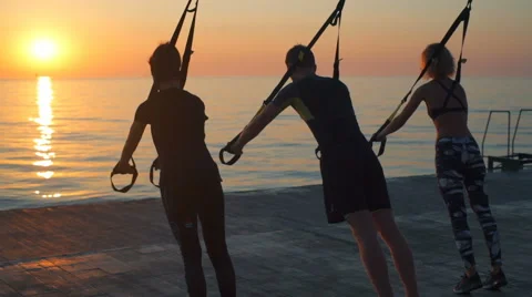Silhouettes of sportive people training arms with trx at sunrise. Slow motion. Stock Footage