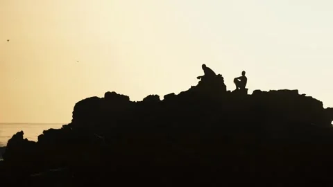 Silhouettes of two people on rocks at sunset: golden hour ocean view, peaceful c Stock Footage 309912277