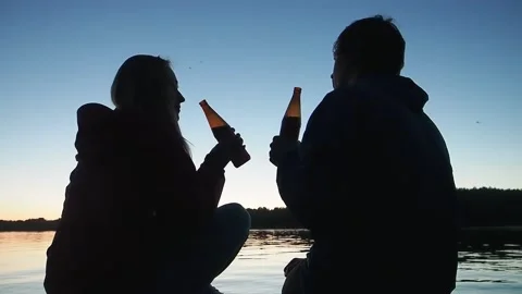 Silhouettes of young couple clinking dark bottles of delicious beer sitting near Stock Footage 153319166
