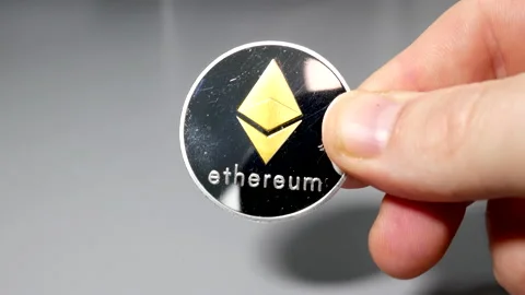 Silver and ethereum bitcoin coin held between fingers in hand on gey table Stock Footage 161376322