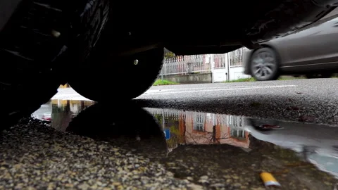 A silver car passing on the road is reflected in the puddle below the parked car Vídeo Stock 221909889