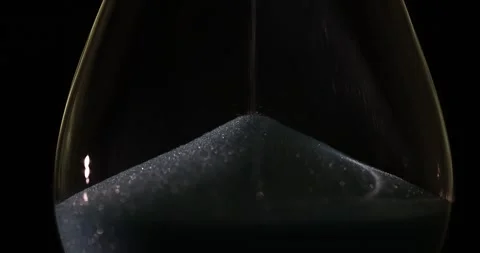 Silver glittery sand cascades through hourglass on black Video stock 281377232