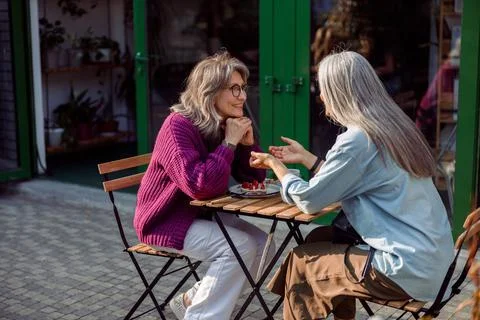 Silver haired woman tells story to mature friend sitting at table outdoors Stock Photos