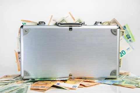 A silver suitcase is sitting on a white surface surrounded by euro and US d.. Stock Photos
