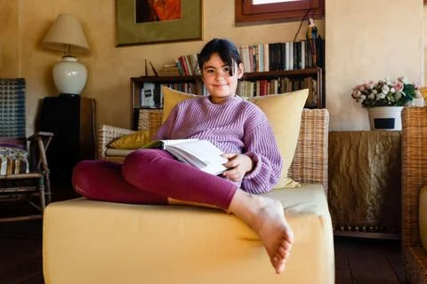 Similing girl in lilac pullover reading a book in yellow armchair 스톡 사진