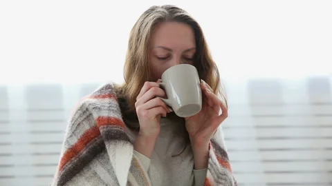 A simple woman drinks tea wrapped in a warm woolen blanket 動画素材 89760865