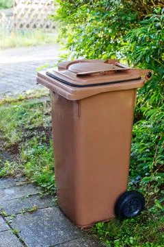 Single brown wheelie bin standing outdoors on a stone path next to green bush Stock Photos
