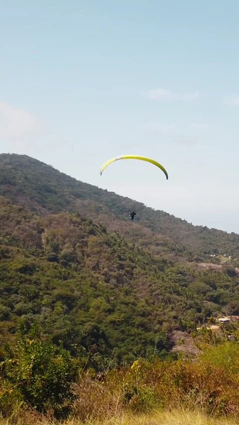 Single paraglider with yellow canopy gliding over green hills and vegetation Stockbeeldmateriaal 332824405