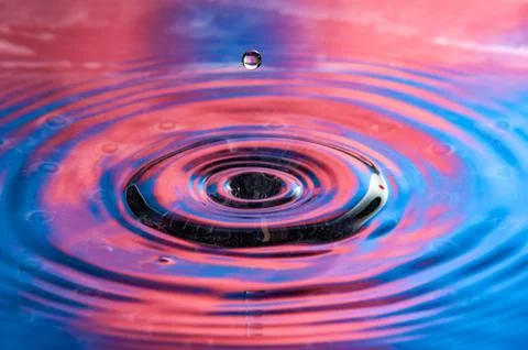 Single water drop falling towards a vibrant red and blue water splash Stock Photos