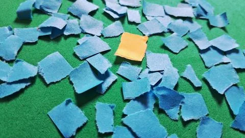 Single yellow paper square surrounded by many small blue pieces Stock Photos