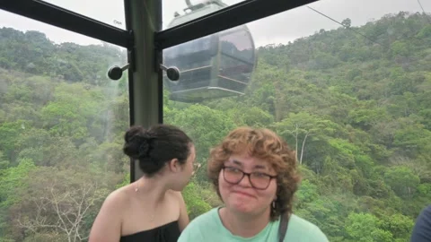 Sisters laughing in cable car in motion above lush hill. 動画素材 330215256