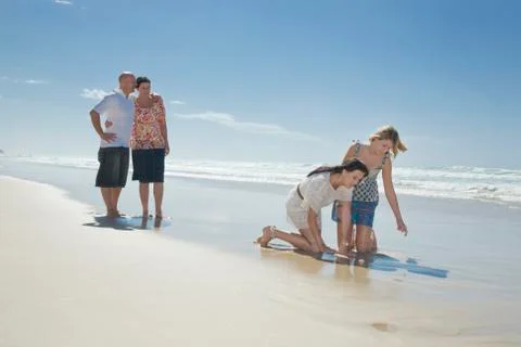 Sisters looking at shell on beach with parents in background Stock Photos