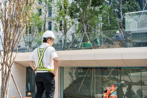 Site engineer standing with hands on waist while observing workers and plan.. Stock Photos