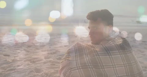 Sitting on beach, man wrapped in blanket with animation of bokeh lights Video stock 305285044