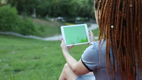 Sitting on the grass in a park a girl with red dreadlocks uses a tablet, lame Stock Footage 134913341