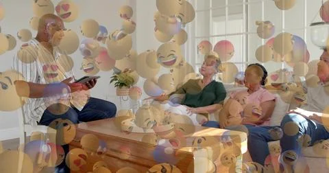 Sitting group of four holding tablet around wood table, floating emoji overlay Stock Photos